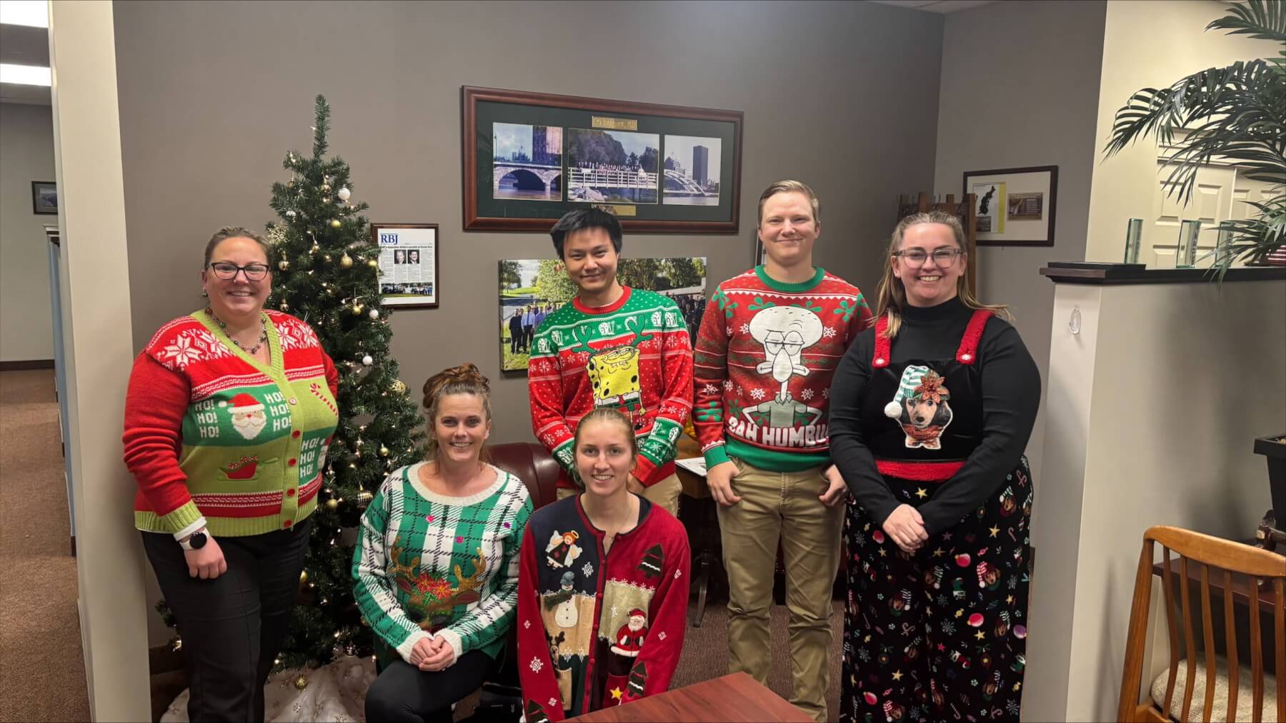 Office team in festive Christmas sweaters by a decorated tree, celebrating holiday spirit.