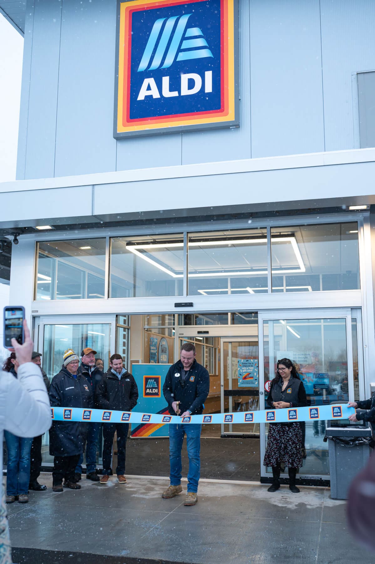 Aldi grand opening with staff cutting ribbon outside new store in snowy weather.