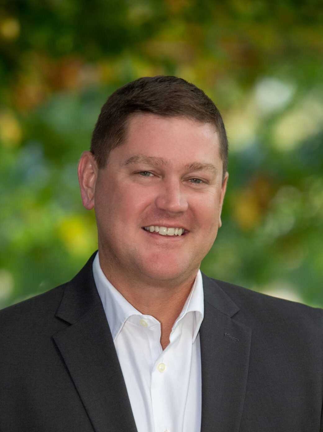 Professional headshot of man standing outdoors with a blurred green background.
