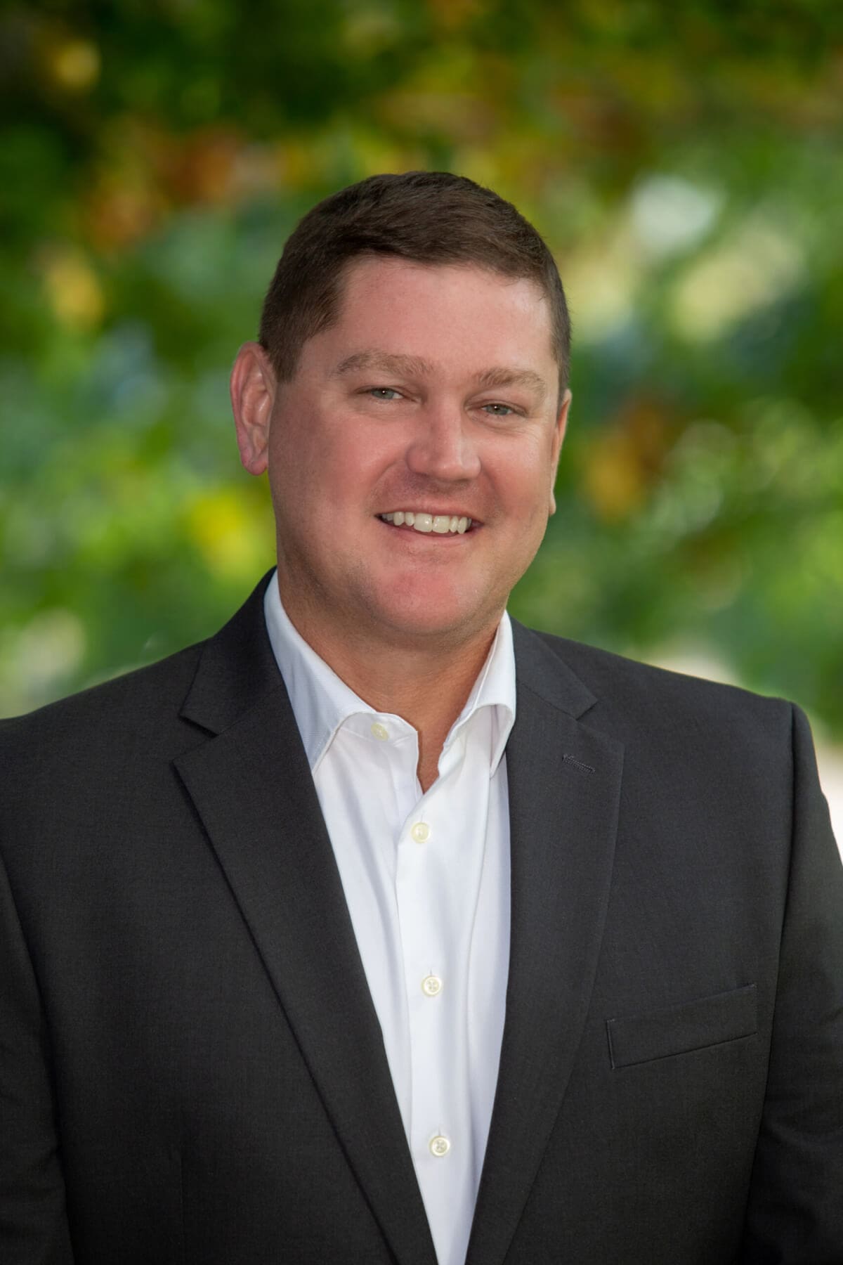 Professional headshot of man standing outdoors with a blurred green background.