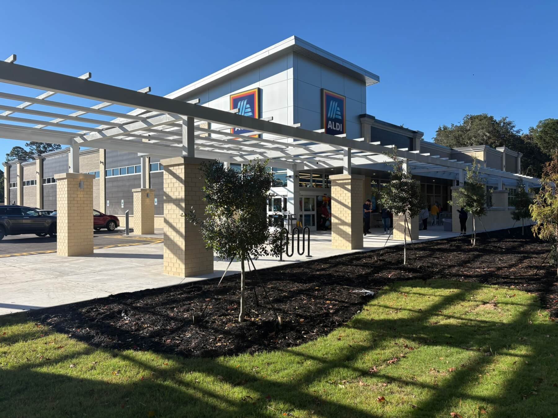 Modern exterior of an ALDI supermarket under clear blue sky, featuring ample parking and landscaped greenery.