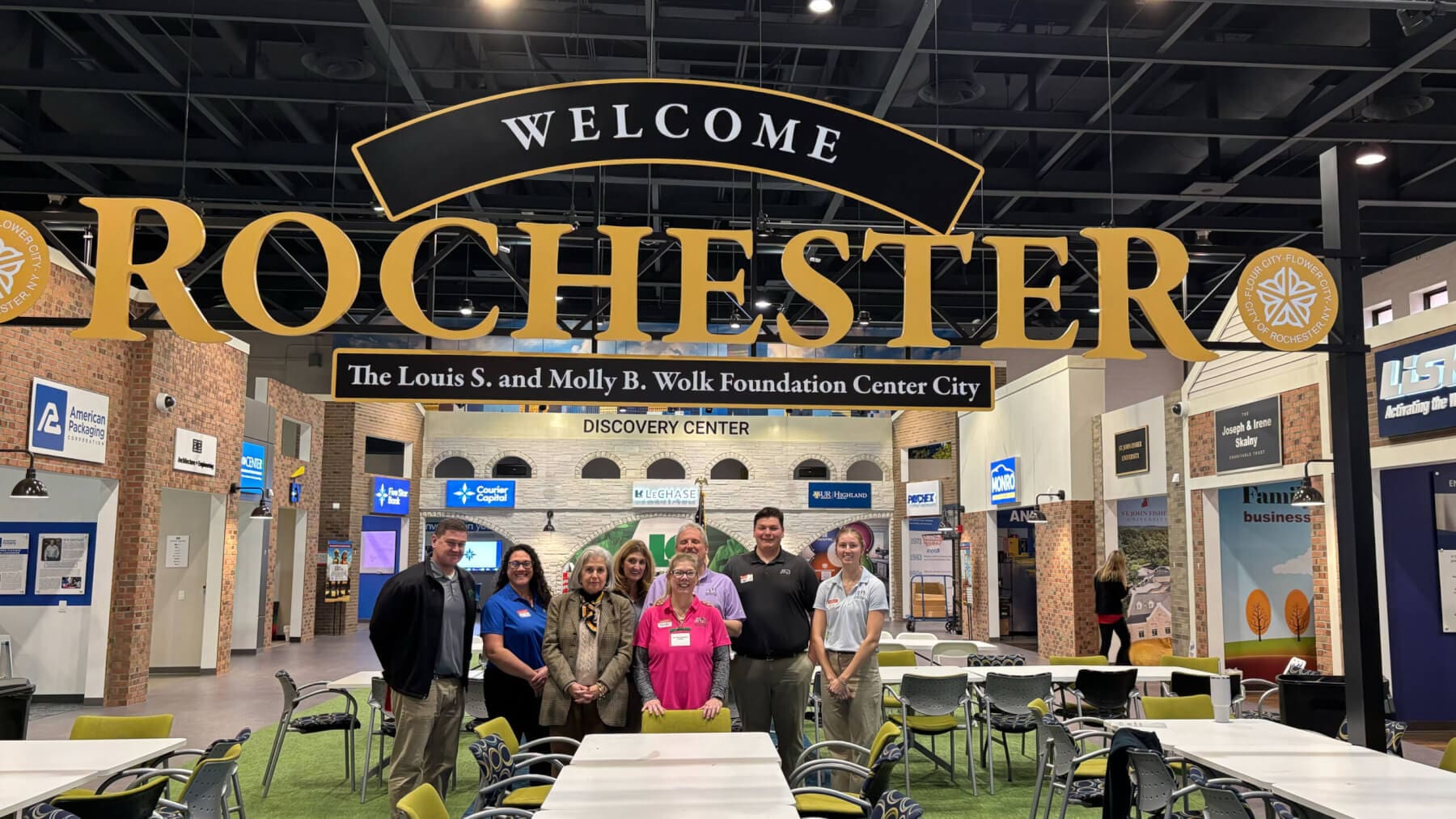 Group poses under Welcome Rochester sign at a discovery center, showcasing local Rochester businesses and culture.