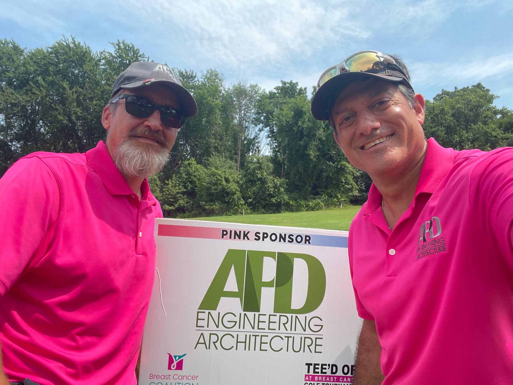 Two smiling men in pink shirts support Breast Cancer Coalition at APD Engineering & Architecture golf event.