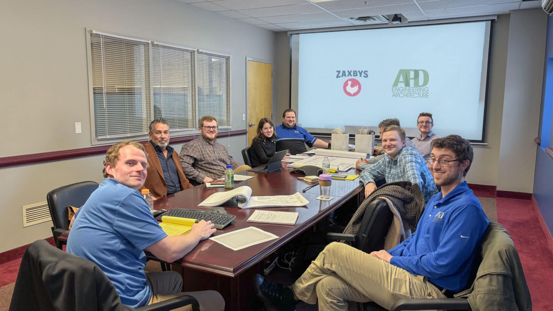 Team meeting in conference room with Zaxby's and APD logos on screen, discussing engineering plans.