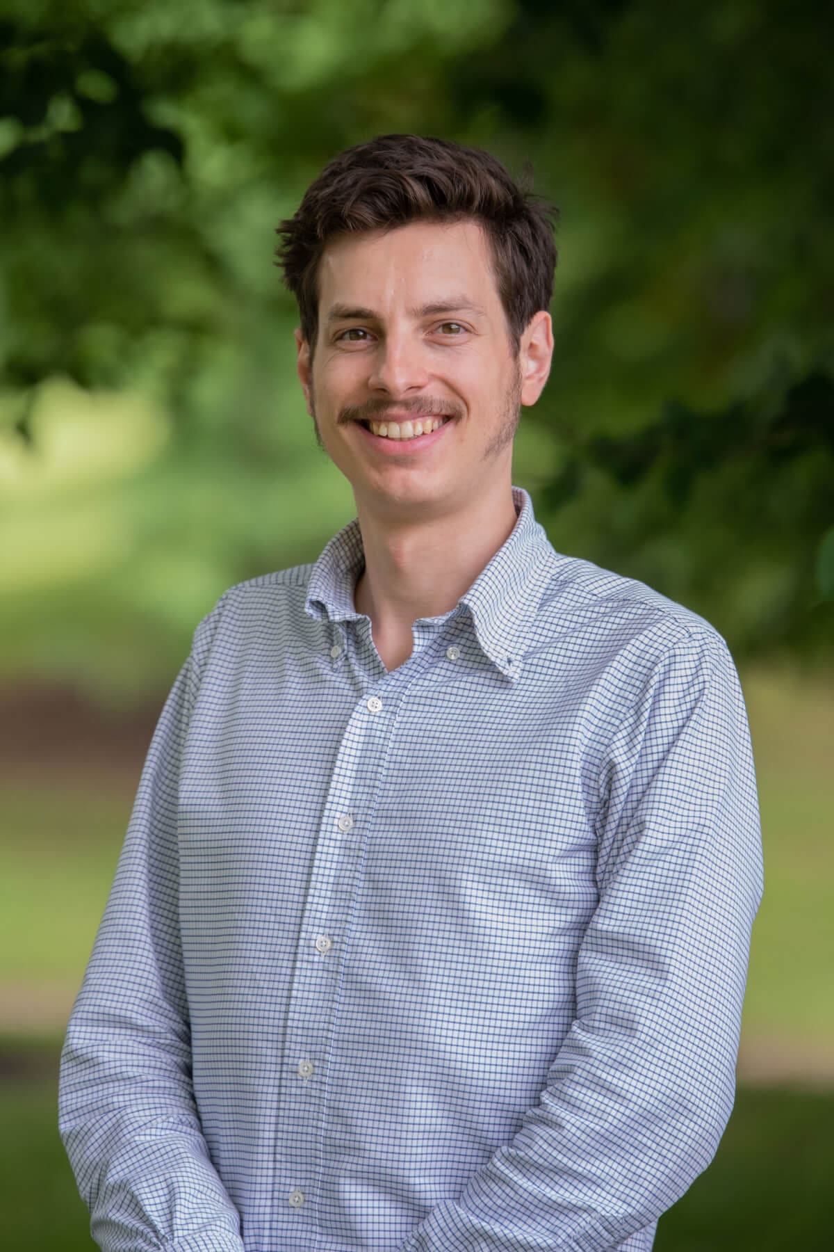 Professional headshot of man standing outdoors with a blurred green background.