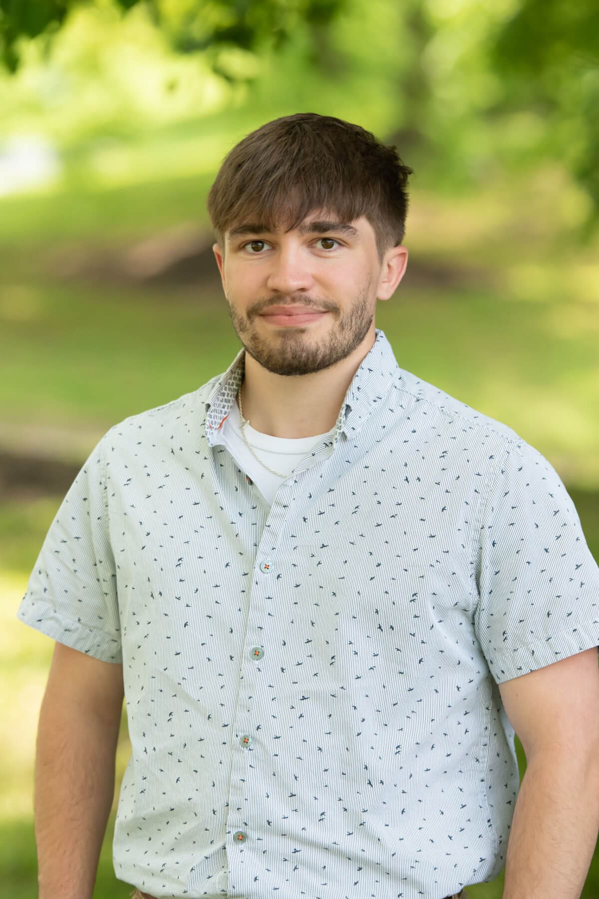 Professional headshot of man standing outdoors with a blurred green background.