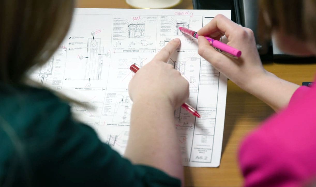 Persons reviewing architectural blueprint, pointing and annotating with pens on a desk, highlighting design details.