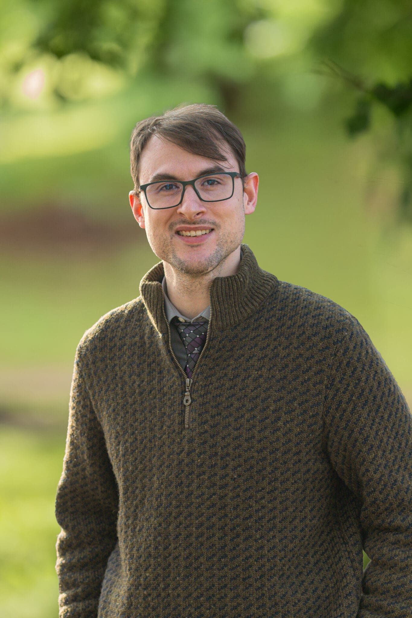 Professional headshot of man standing outdoors with a blurred green background.