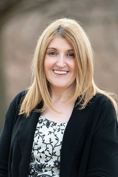 Professional headshot of woman standing outdoors with a blurred brown background.