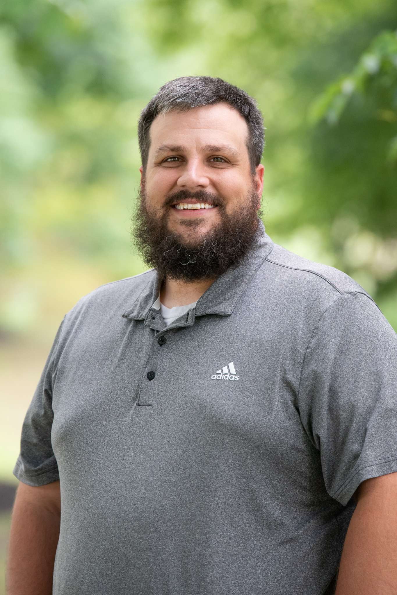 Professional headshot of man standing outdoors with a blurred green background.