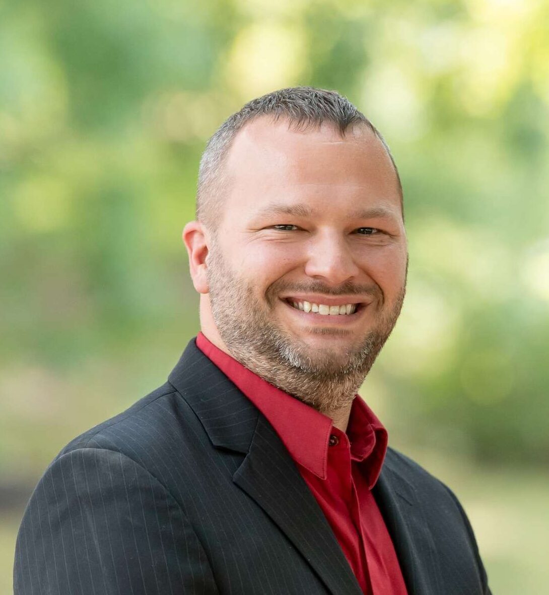 Professional headshot of man standing outdoors with a blurred green background.