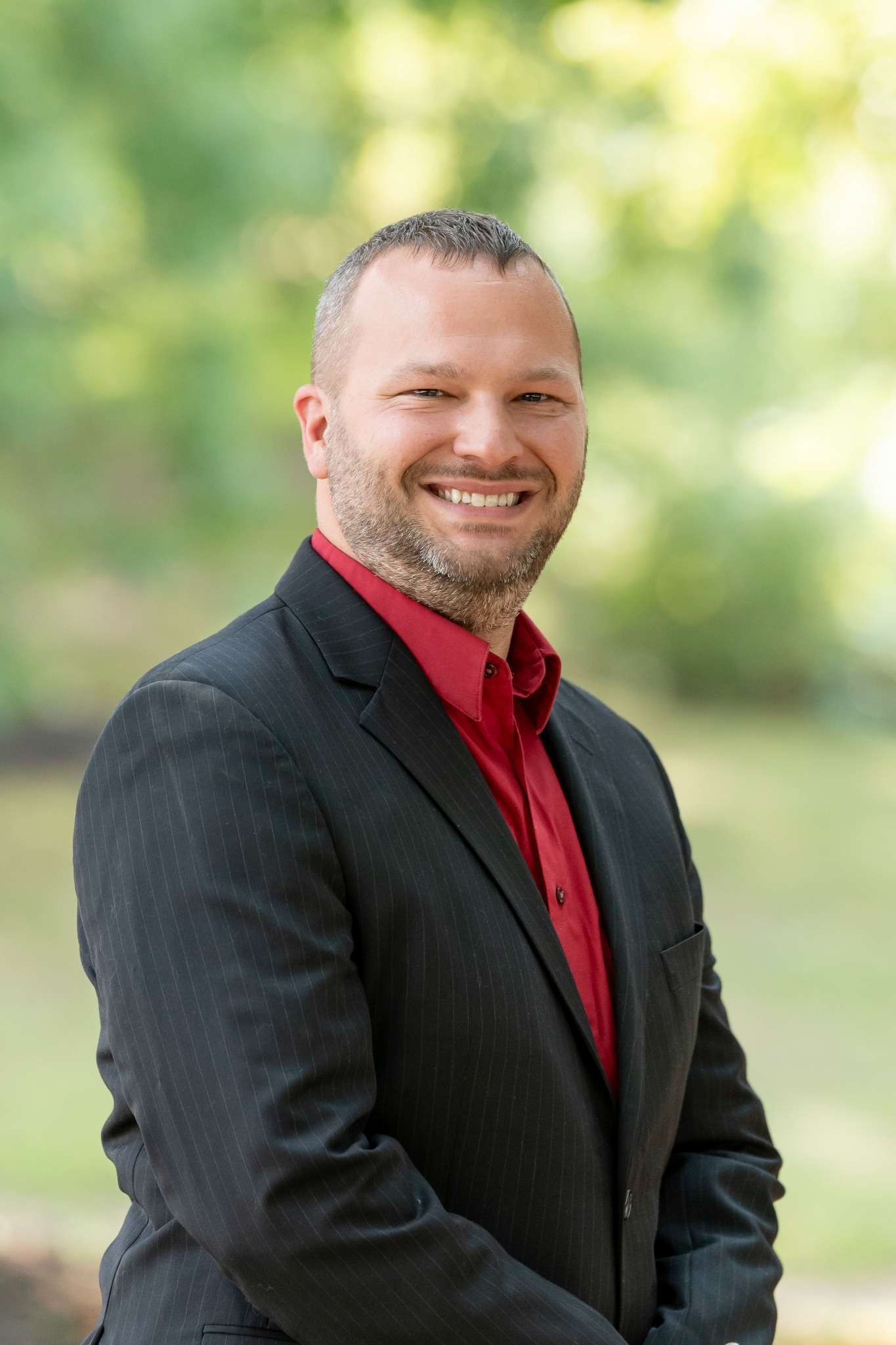 Professional headshot of man standing outdoors with a blurred green background.