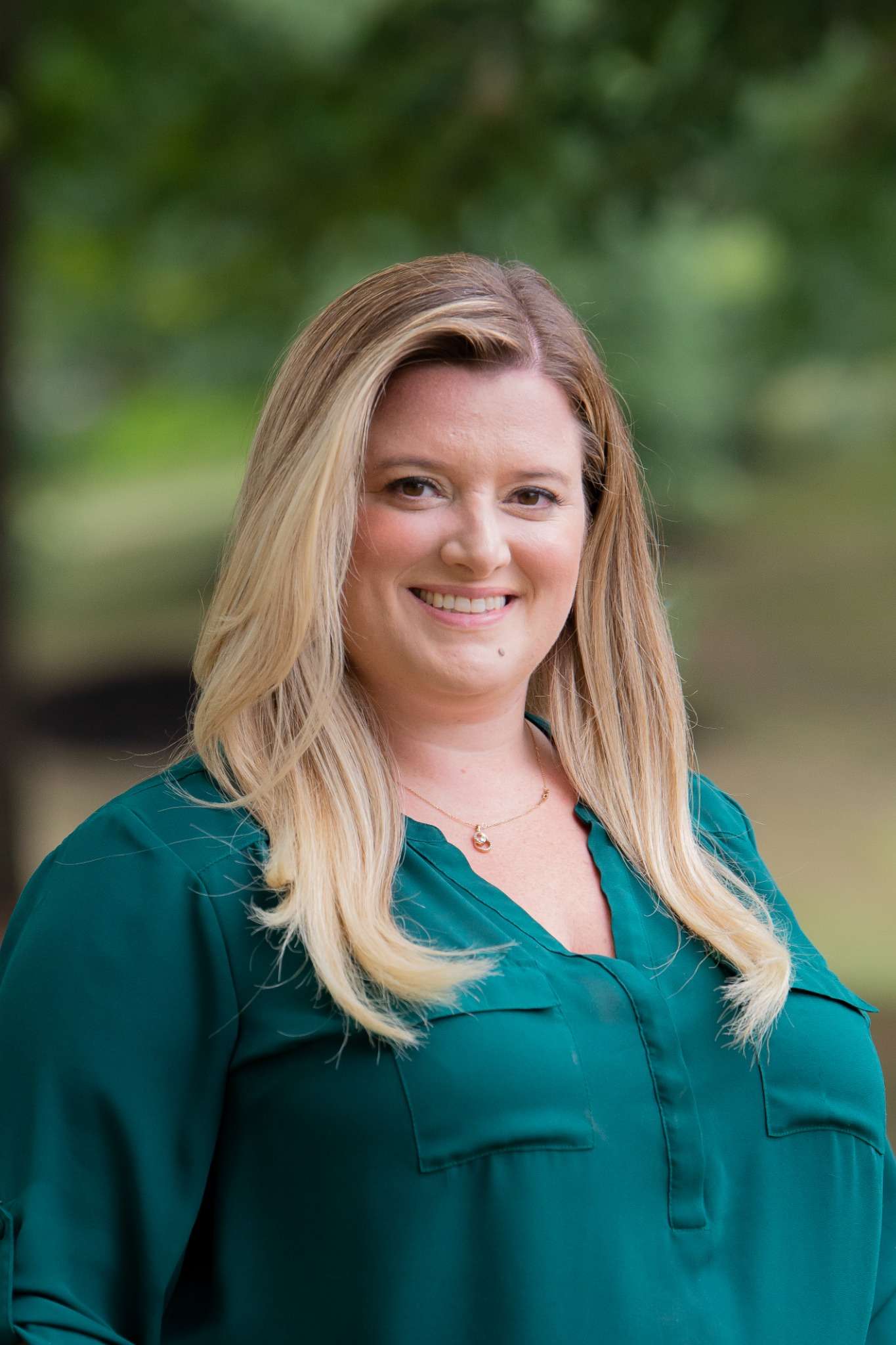 Professional headshot of woman standing outdoors with a blurred green background.