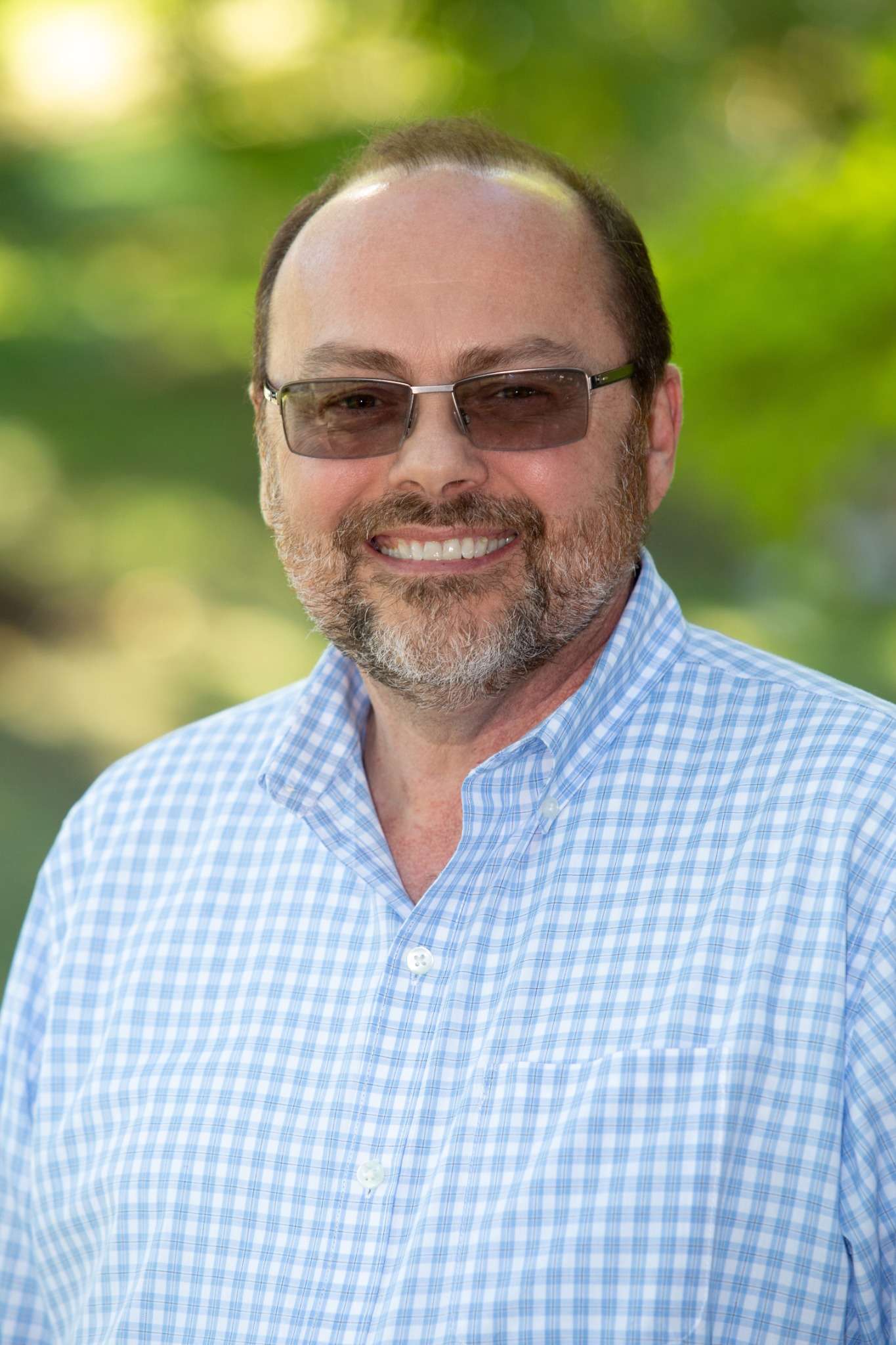 Professional headshot of man standing outdoors with a blurred green background.
