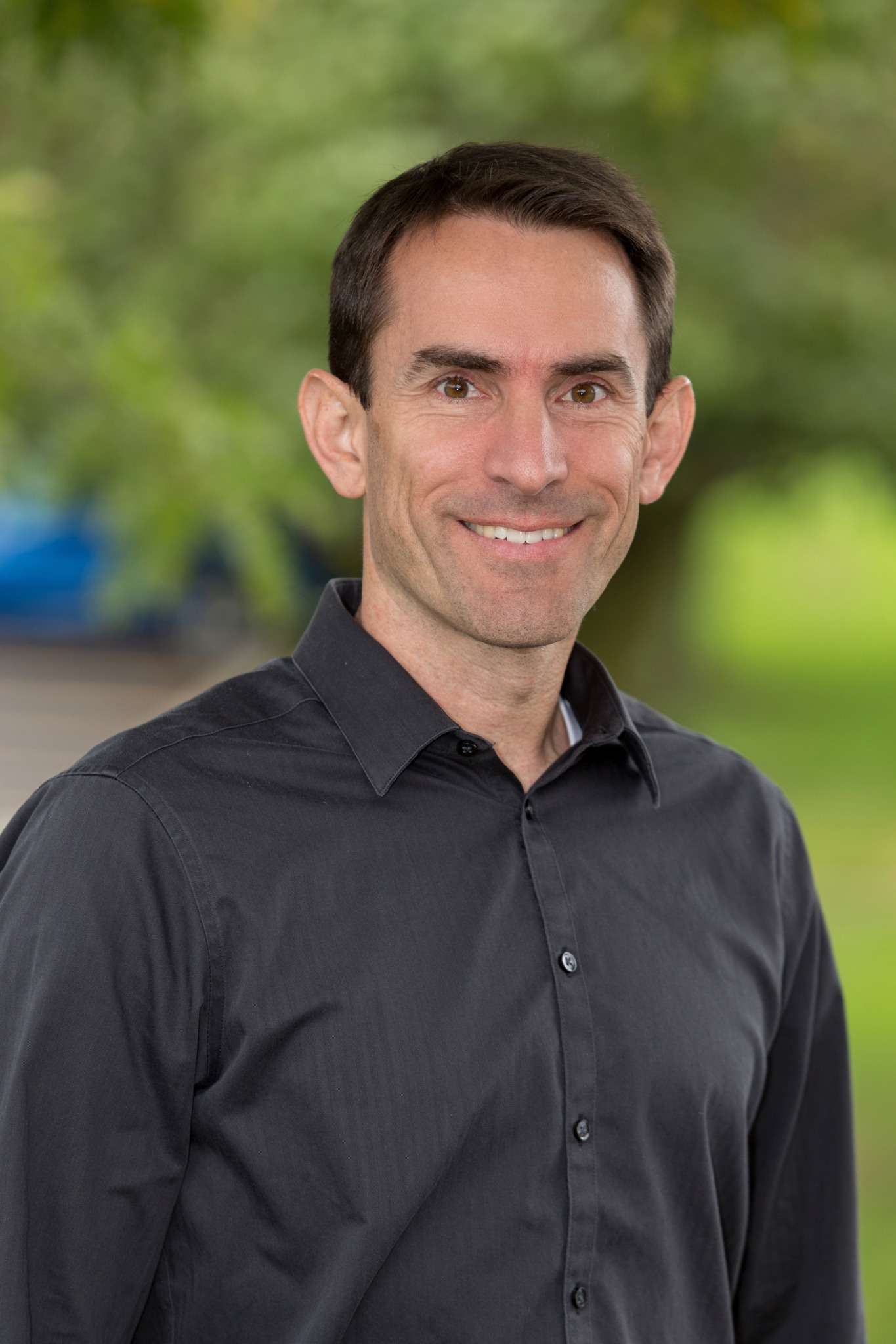Professional headshot of man standing outdoors with a blurred green background.