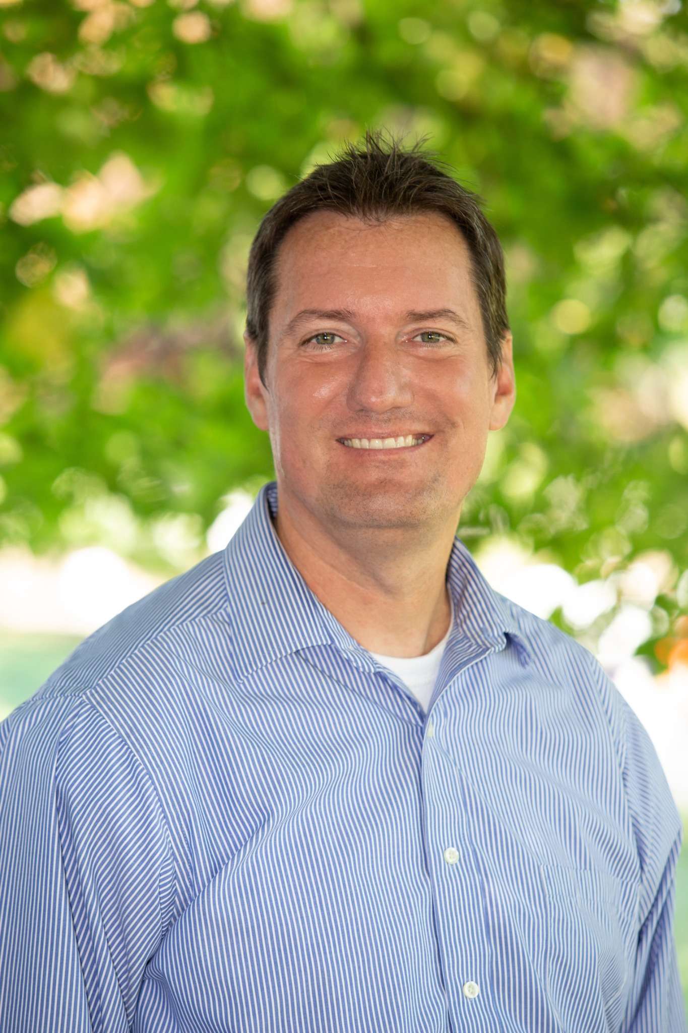 Professional headshot of man standing outdoors with a blurred green background.
