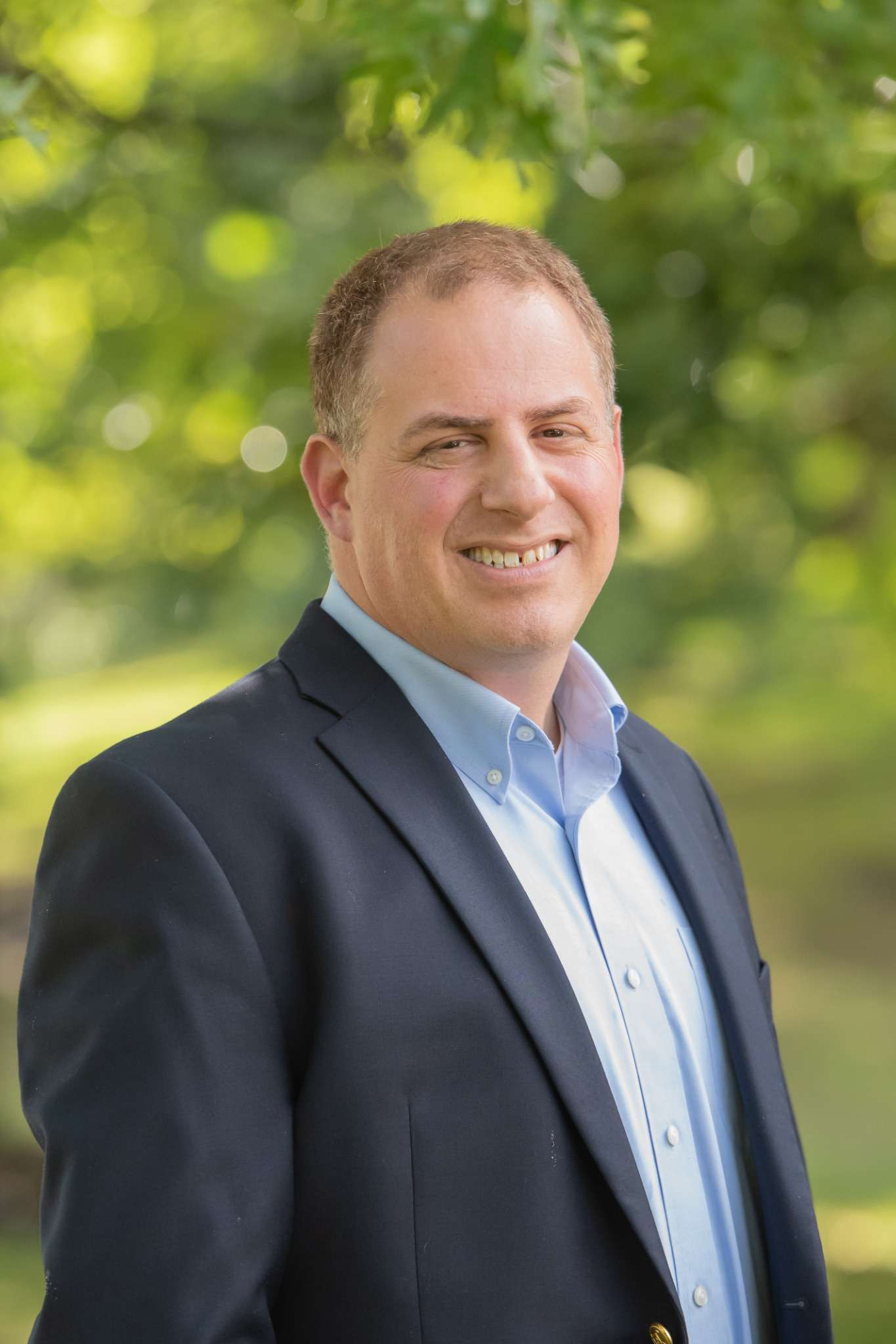 Professional headshot of man standing outdoors with a blurred green background.