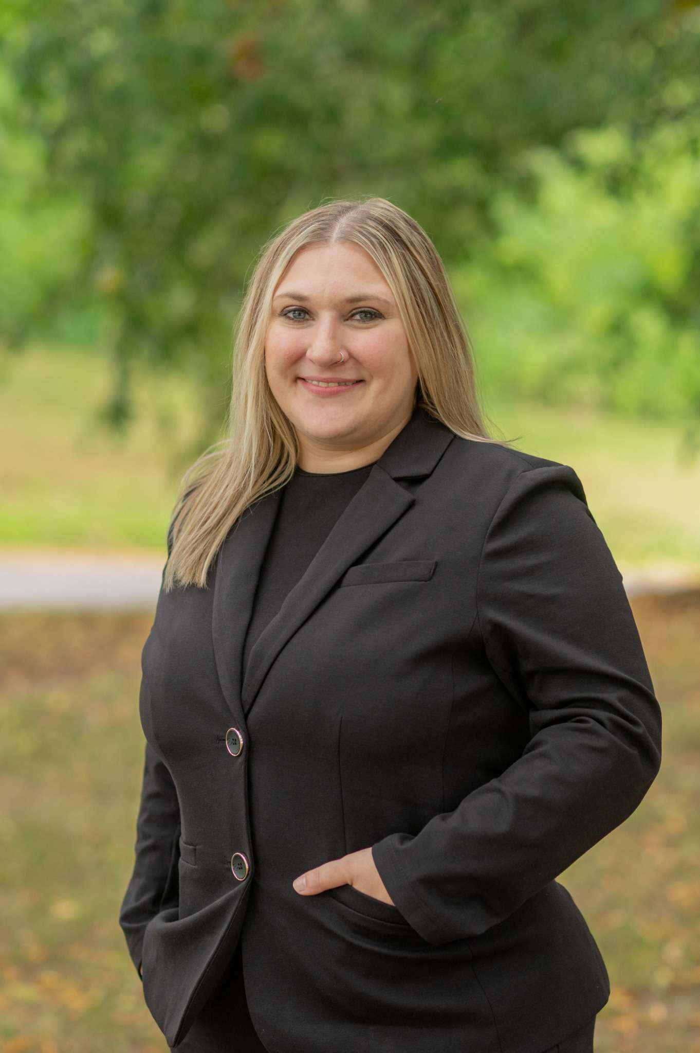 A professional woman in a black blazer smiles in a park setting.