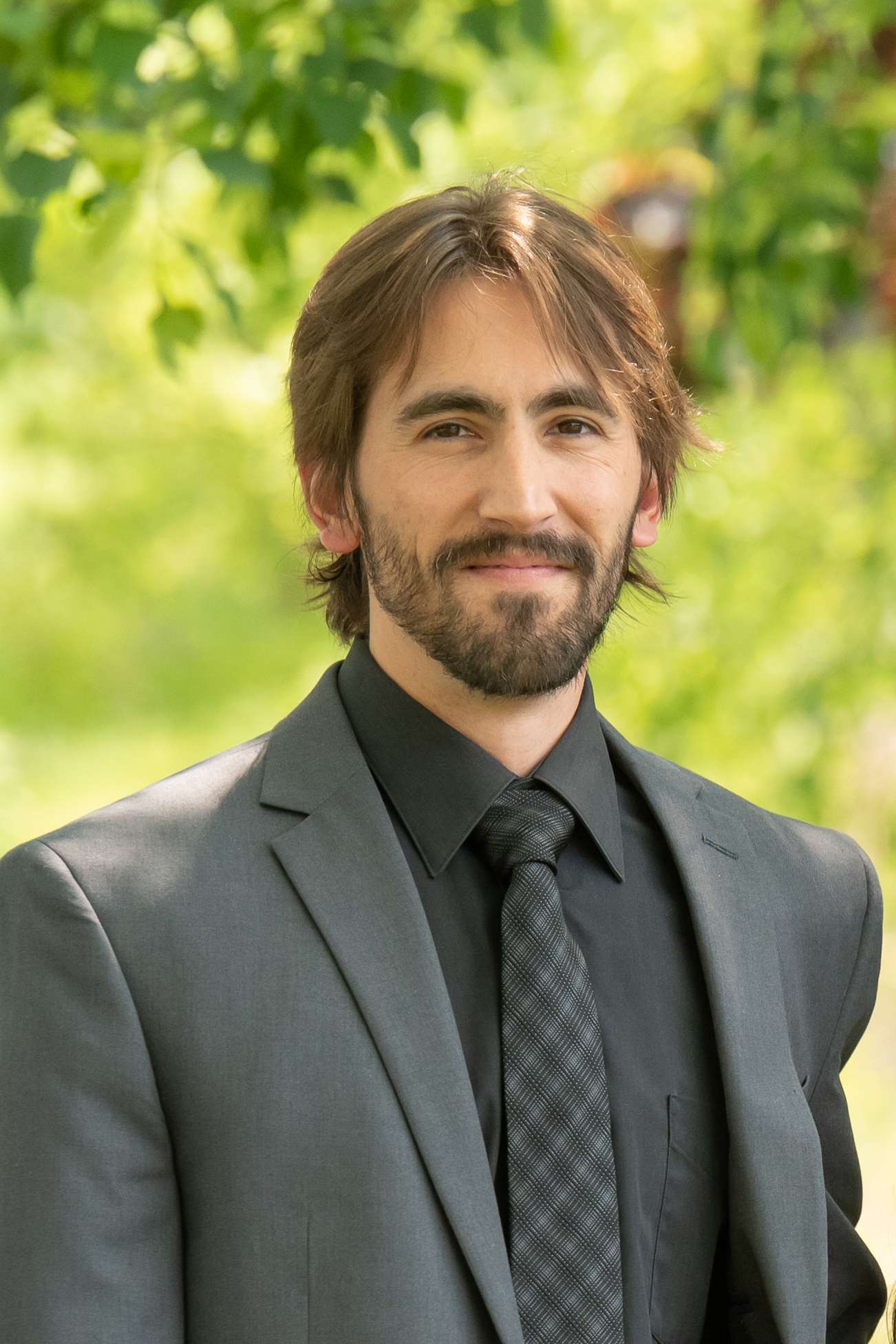 Professional headshot of man standing outdoors with a blurred green background.