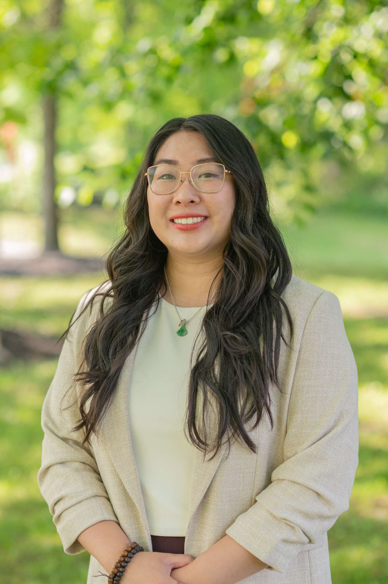 Friendly woman in a beige blazer smiling in a lush park setting.
