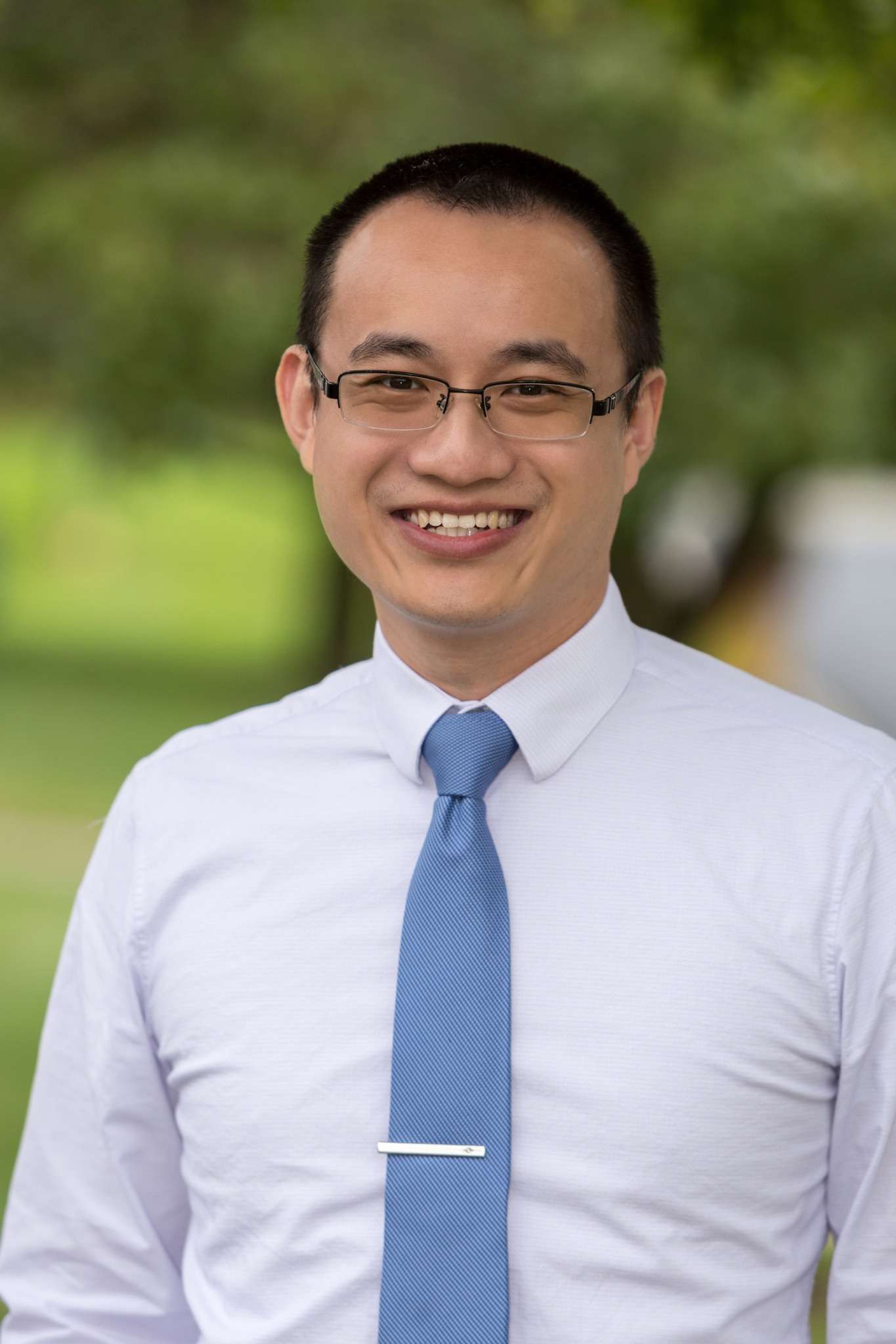 Professional headshot of man standing outdoors with a blurred green background.