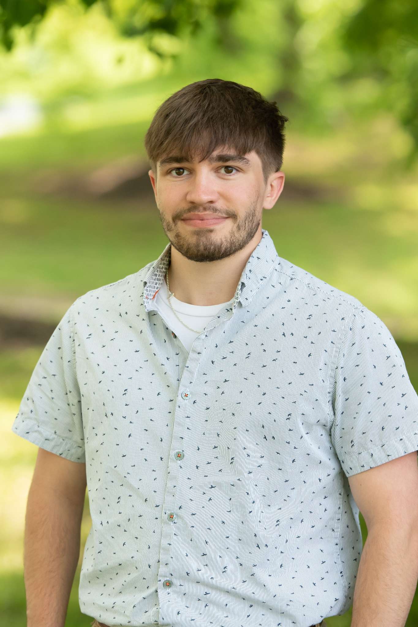 Professional headshot of man standing outdoors with a blurred green background.