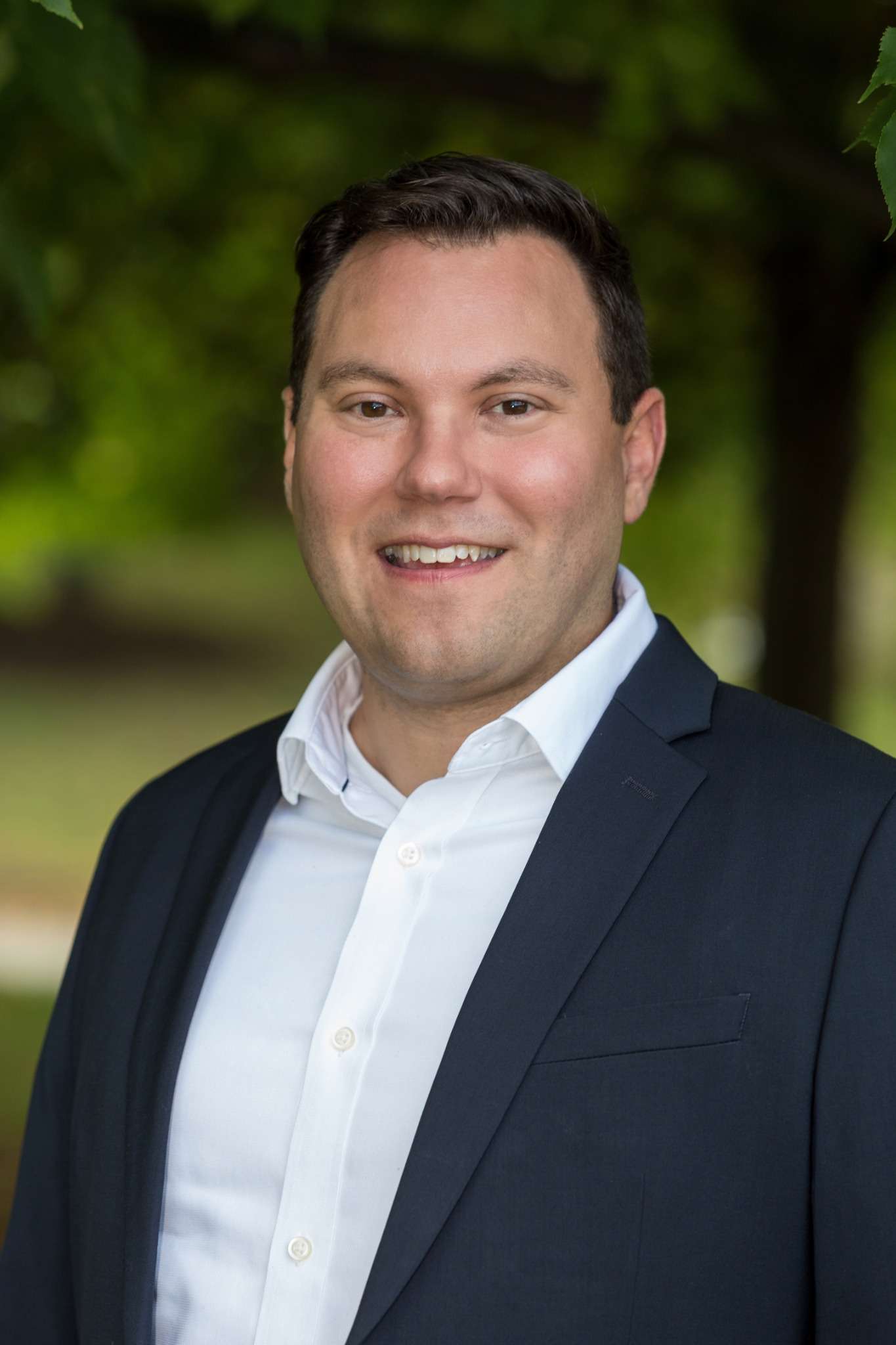 Professional headshot of man standing outdoors with a blurred green background.