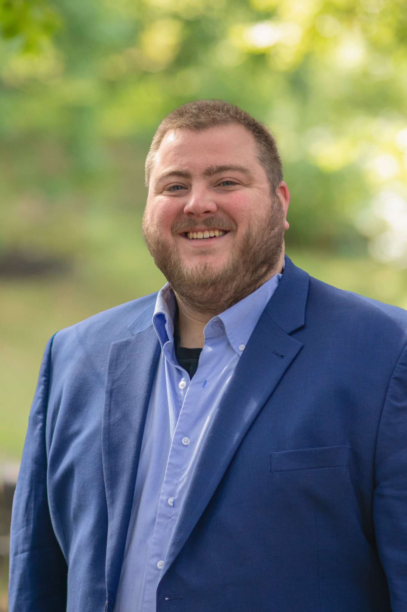 Professional headshot of man standing outdoors with a blurred green background.