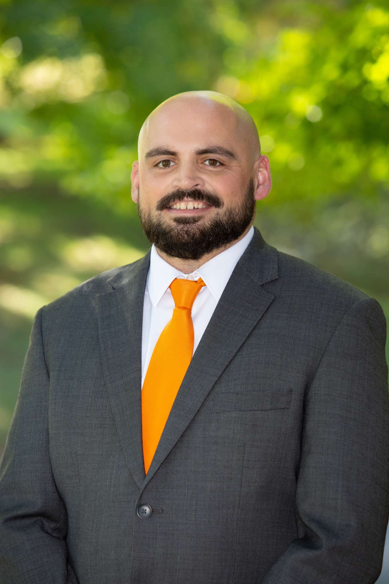 A professional man in a suit with an orange tie, posing outdoors.