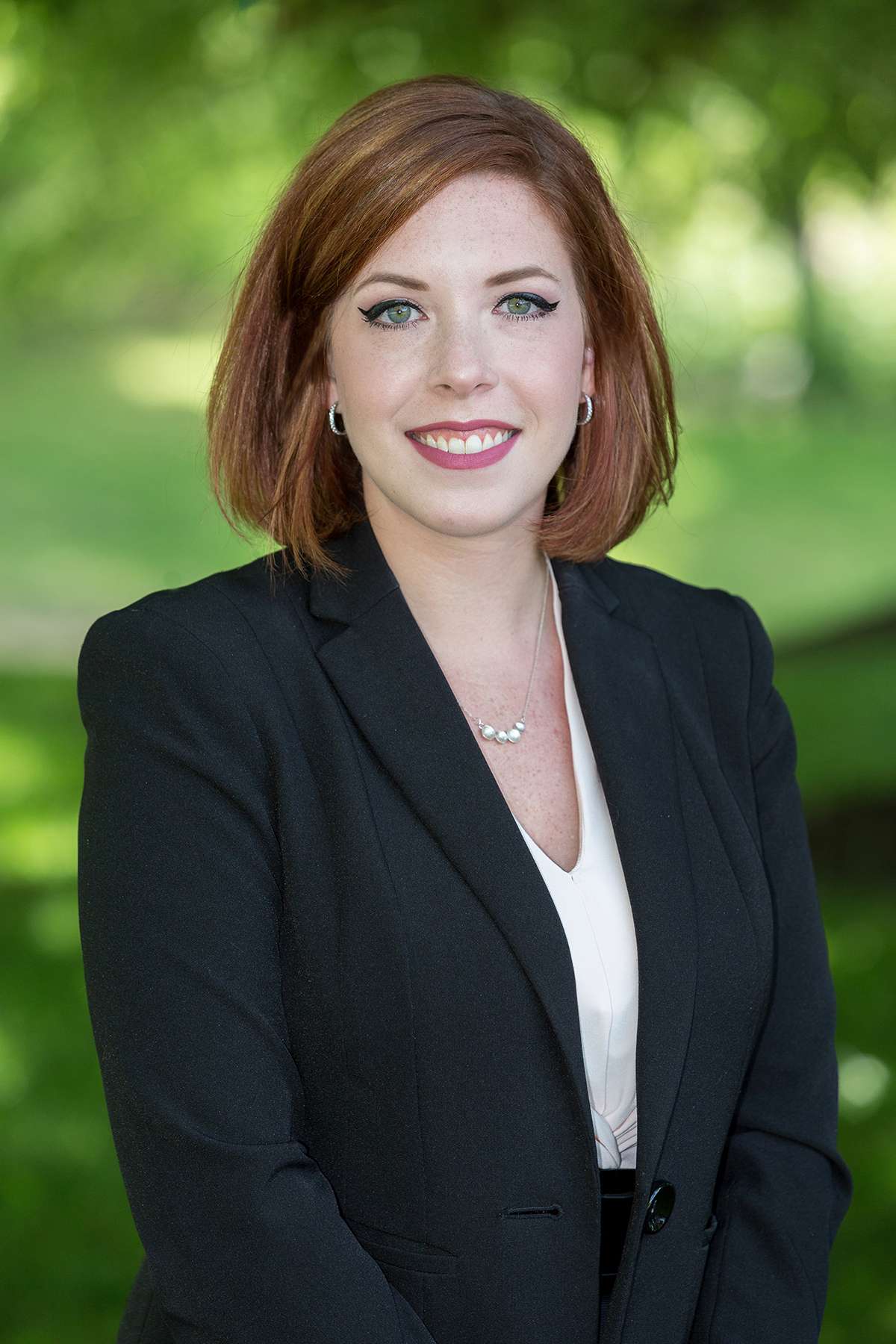Professional headshot of woman standing outdoors with a blurred green background.