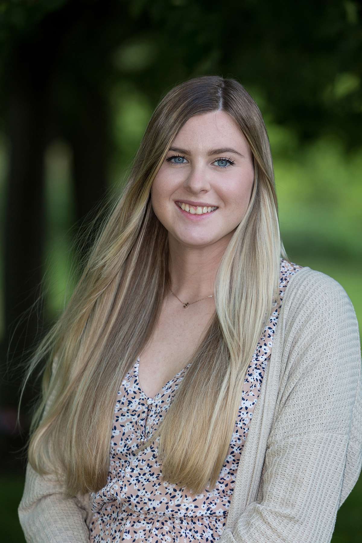 Smiling woman with long blonde hair in a floral dress, outdoors in a leafy park setting.