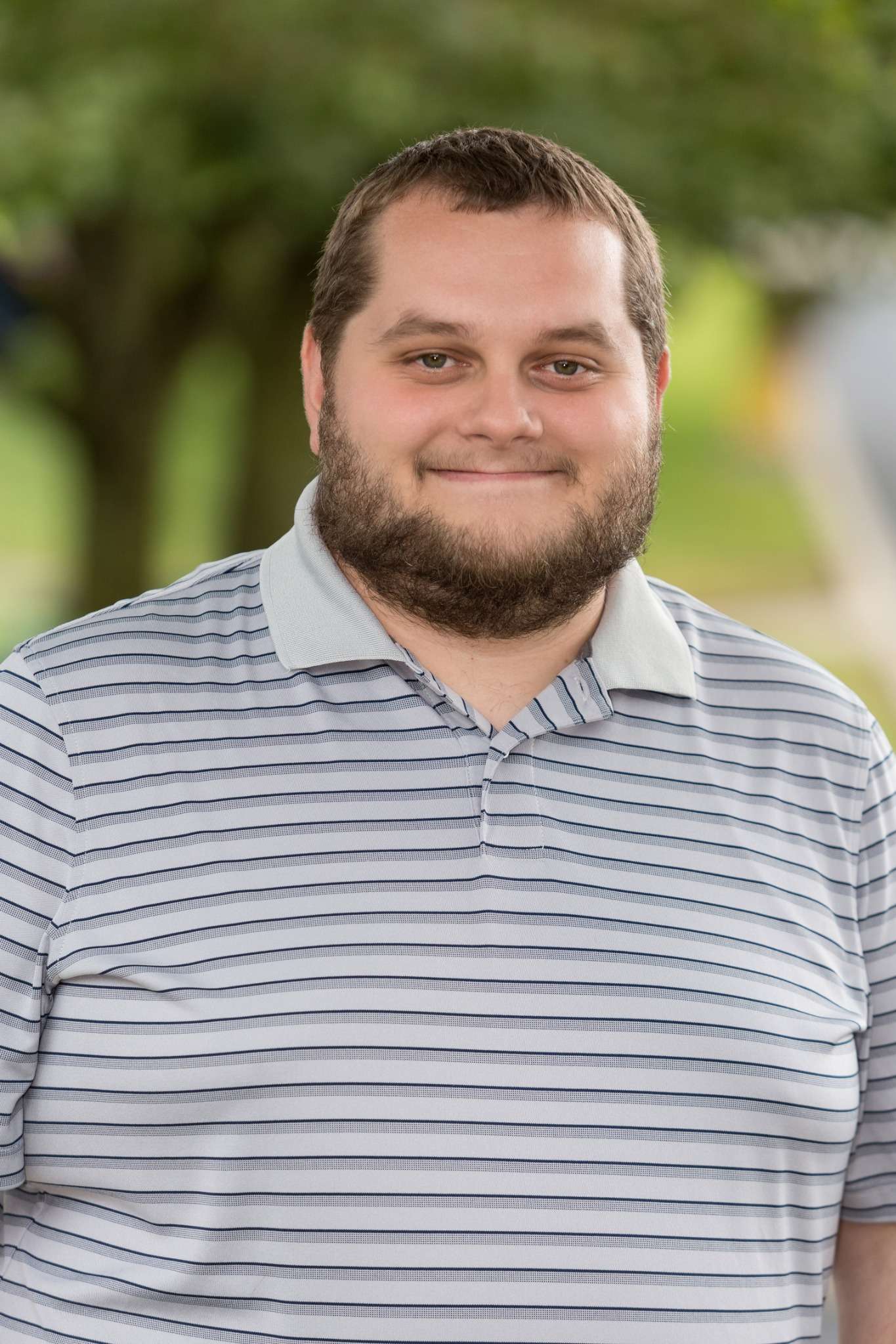 Professional headshot of man standing outdoors with a blurred green background.