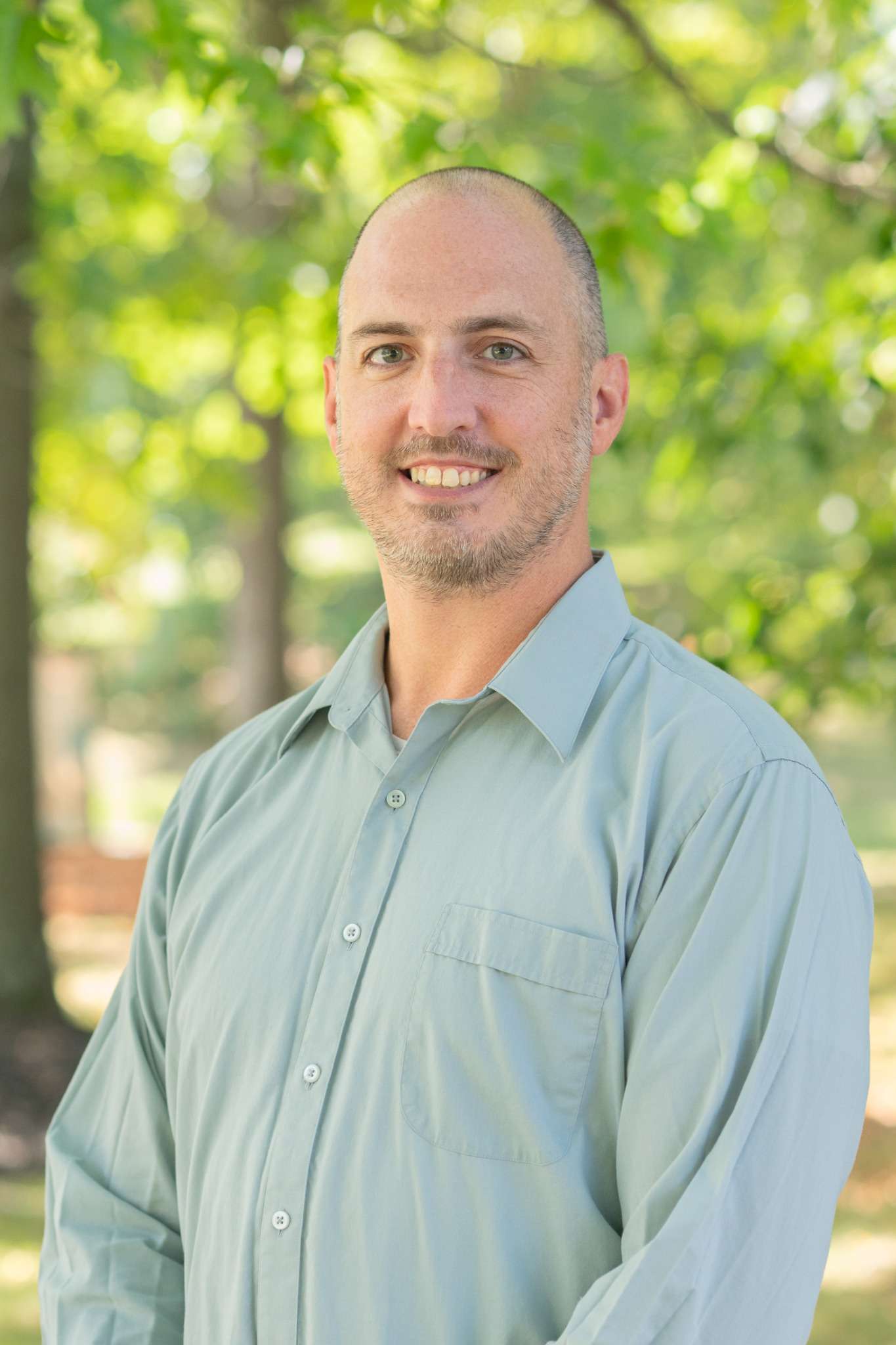 Professional headshot of man standing outdoors with a blurred green background.