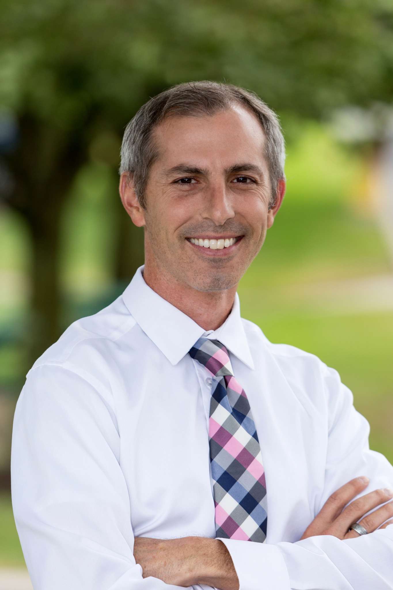 Professional headshot of man standing outdoors with a blurred green background.