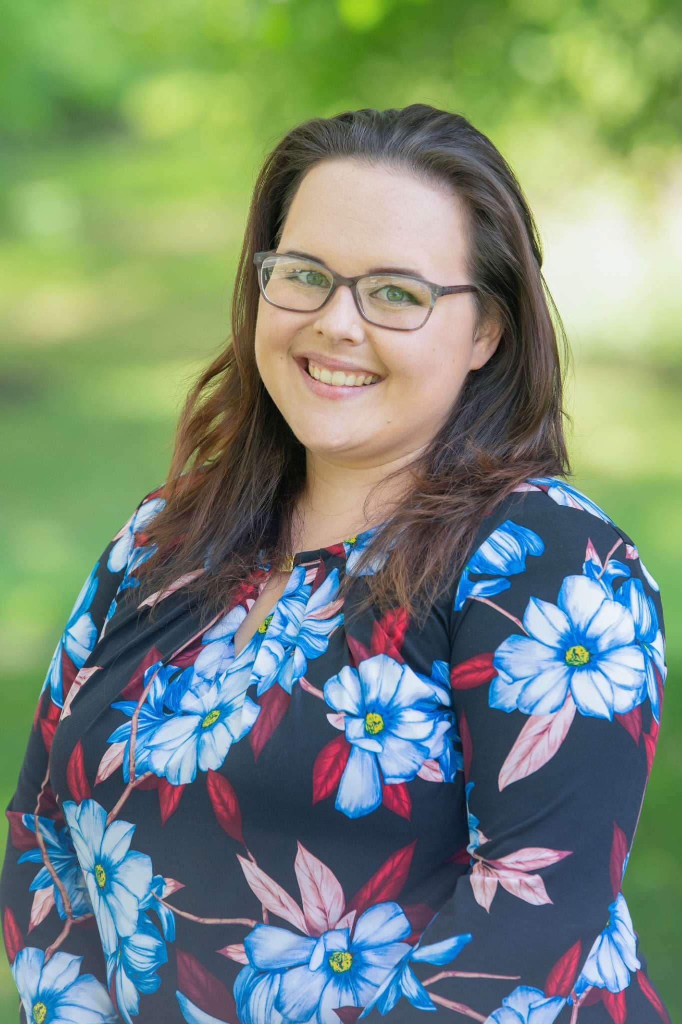 Smiling woman in glasses with blue floral blouse against a blurred greenery background.