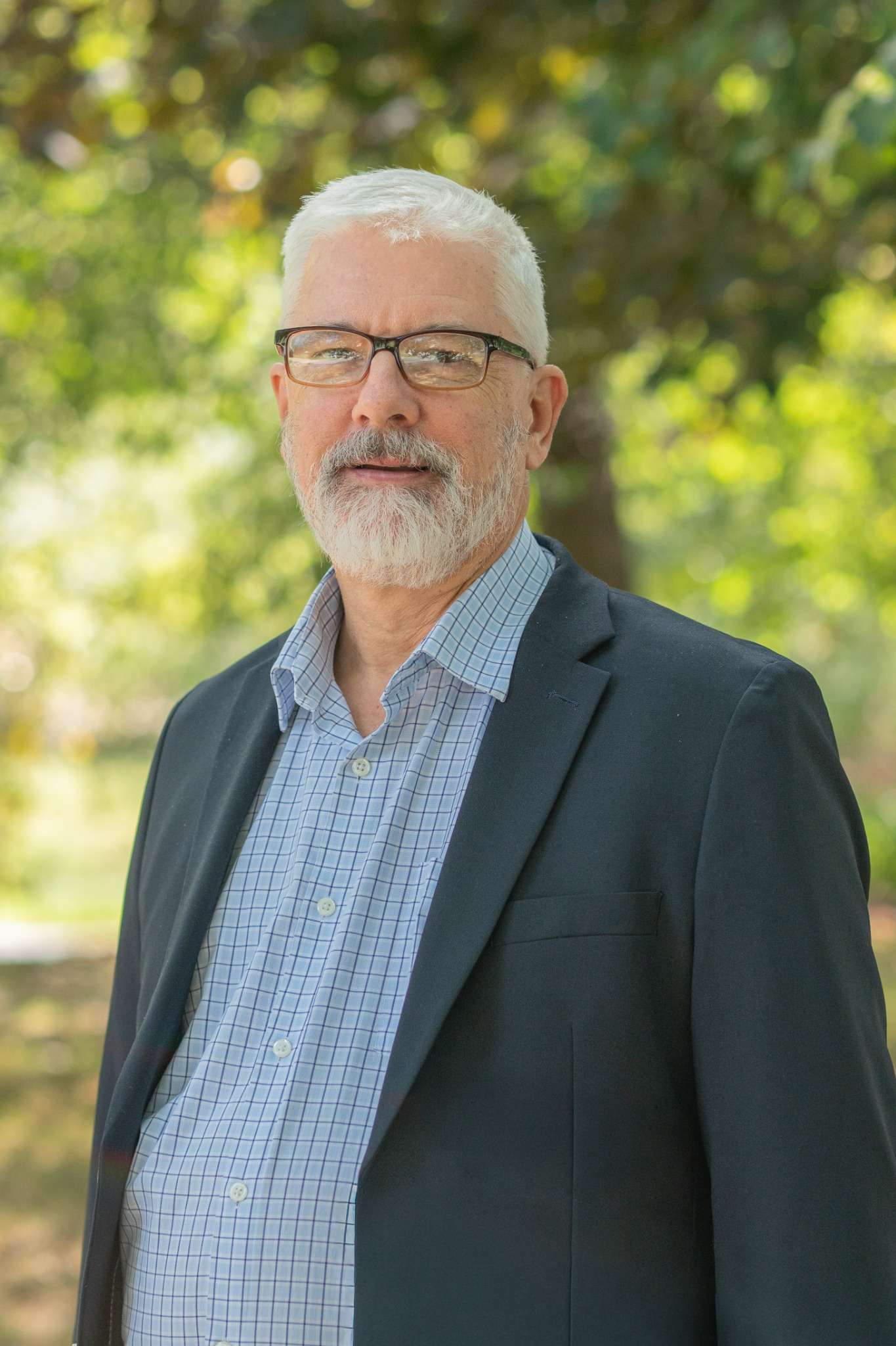 Professional headshot of man standing outdoors with a blurred green background.