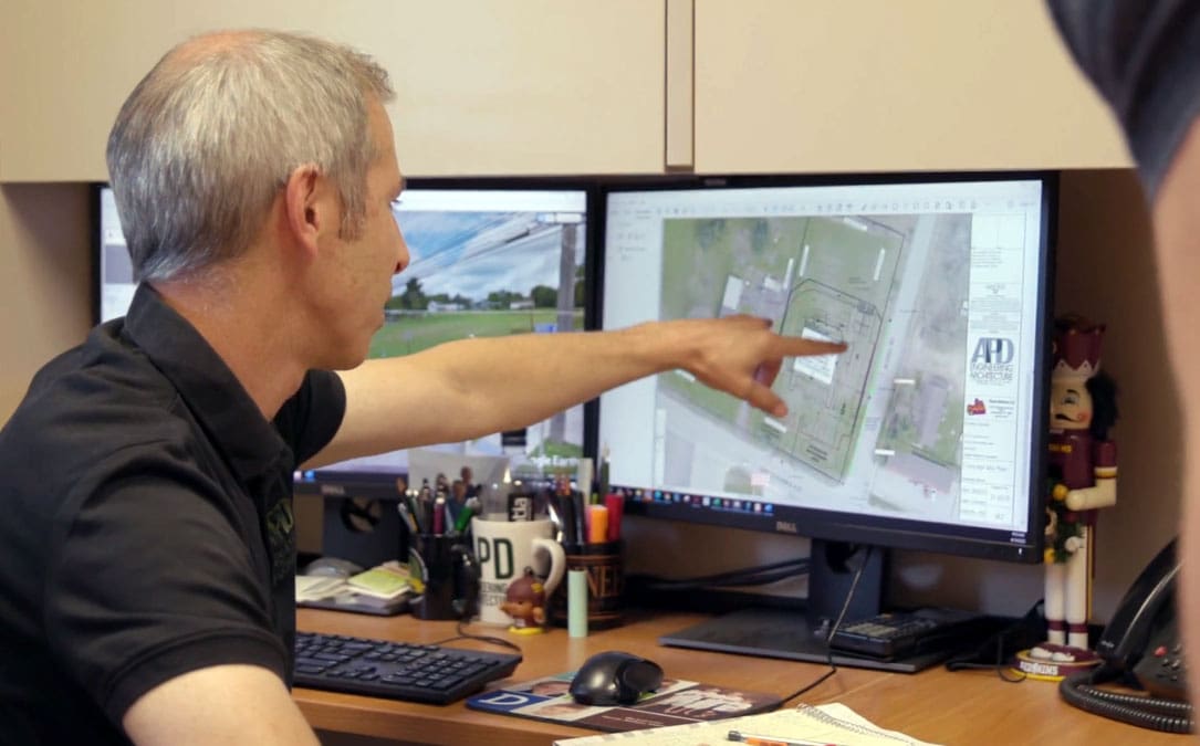Man reviewing architectural plans on dual monitors, engaged in site planning at APD Engineering office.