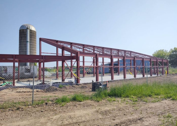 Steel barn under construction on a sunny day, surrounded by green fields and a grain silo in the background.