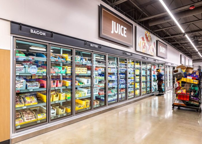 Grocery store aisle with refrigerated sections for bacon, butter, and juice; shopper in the background.