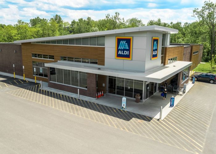 Exterior view of an Aldi grocery store with parking area, set against a backdrop of green trees and a blue sky.