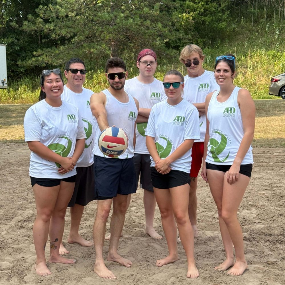 Team in branded shirts stands together on sand, holding a volleyball at an AHD company event.