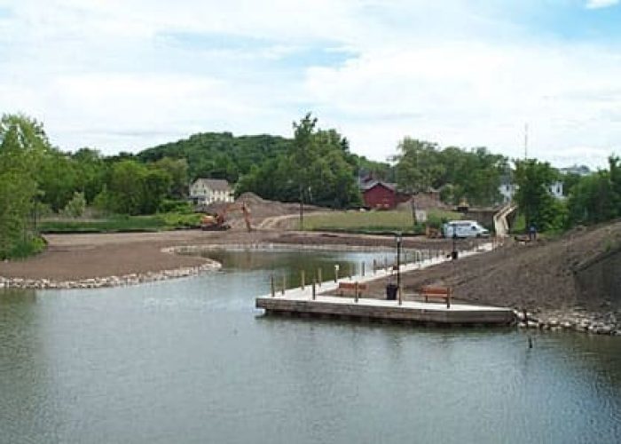 Riverfront park under construction near a bridge, featuring scenic greenery and a small dock.