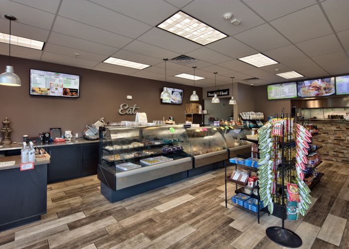 Modern deli interior with display cases, snack racks, and overhead digital menus for customer ordering.