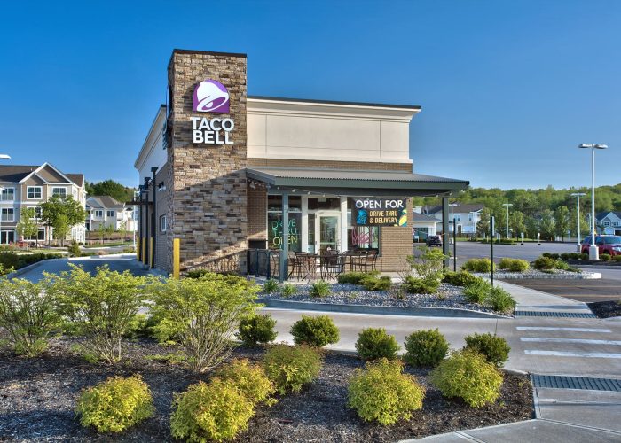 Taco Bell restaurant with drive-thru and delivery options available, set against a clear blue sky.