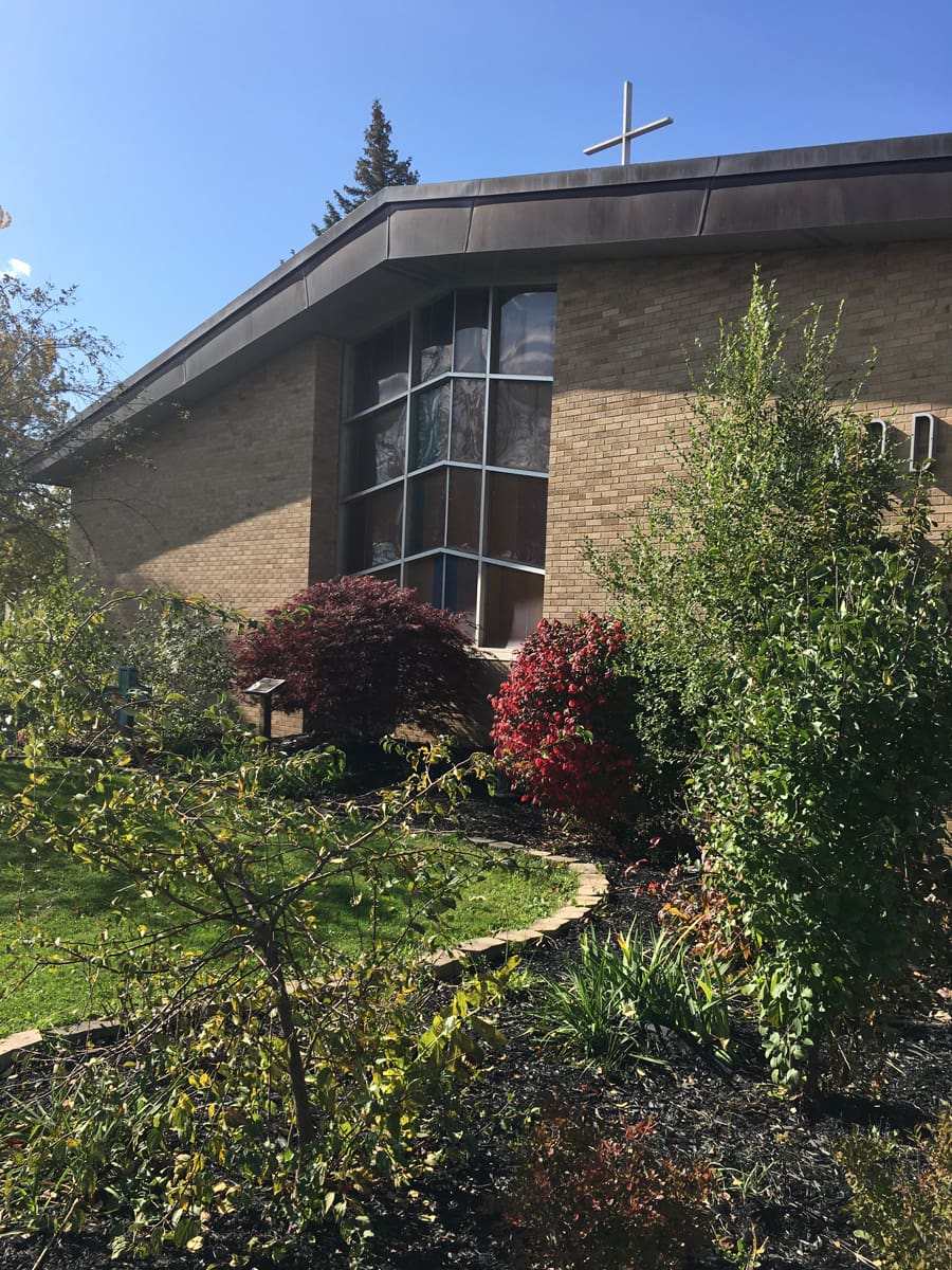 Modern brick church building with cross, surrounded by vibrant garden on a sunny day.