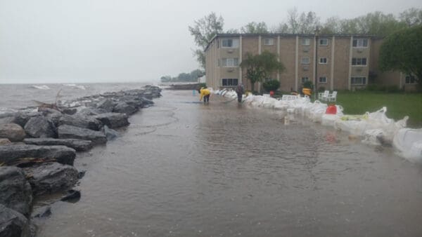 Floodwaters near an apartment building with sandbags and rocks along the shoreline for protection.