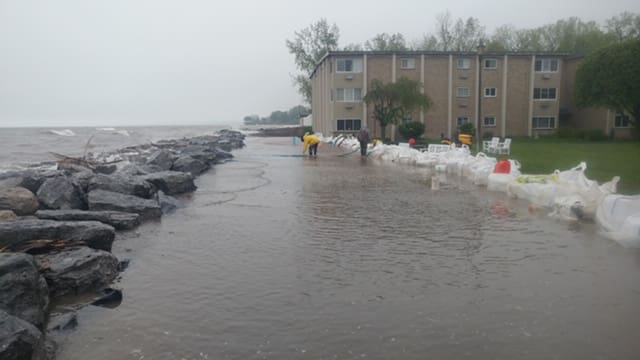 Floodwaters near an apartment building with sandbags and rocks along the shoreline for protection.
